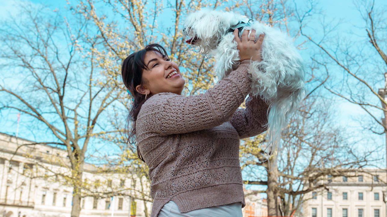 Young woman holding small white dog in the air, smiling, with Waterbury buildings in the background