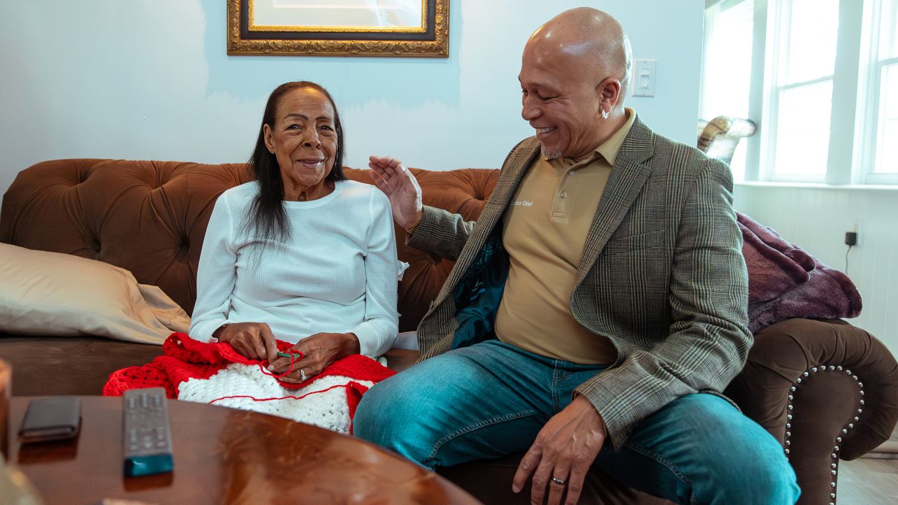 Man sitting with his senior mother on couch inside living room, as she knits