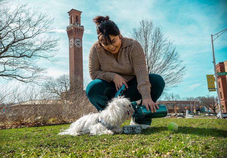 A smiling young woman outdoors, giving water to her dog in a bowl, with the Waterbury Clock Tower in the background