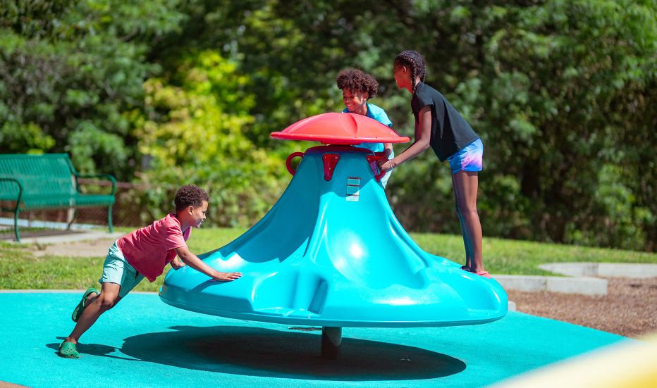 Three kids playing at the playground, with one kid pushing a spinning playground element while the other two kids ride it
